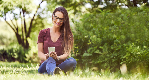 Smiling young woman sitting on ground in a park and using smartphone