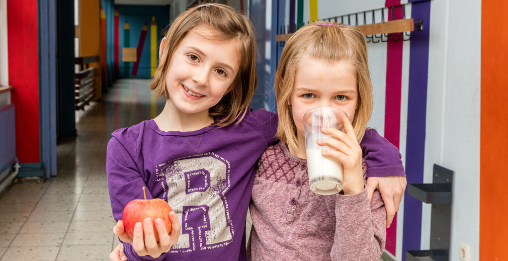 Mädchen im Schulflur mit Milchglas und Apfel.